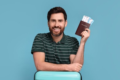 Smiling man with passport, suitcase and tickets on light blue background