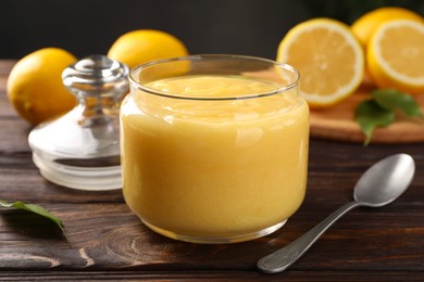 Photo of Delicious lemon curd in glass jar, fresh citrus fruits and spoon on wooden table