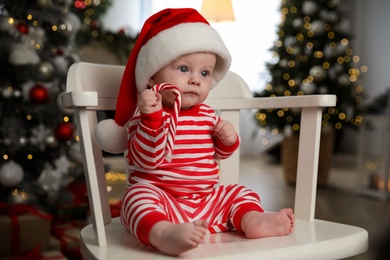 Cute baby in Santa hat and bright Christmas pajamas holding candy cane at home