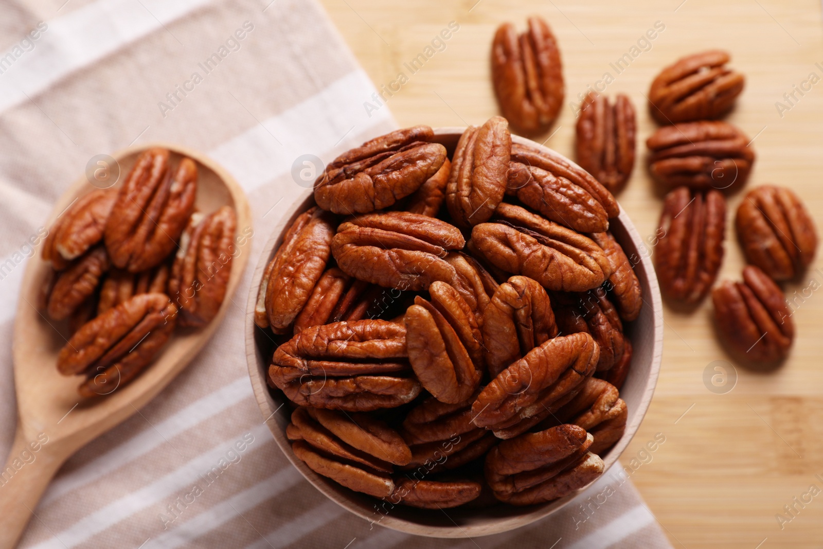Photo of Tasty pecan nuts with bowl, spoon and cloth on wooden table, flat lay