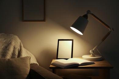 Photo of Stylish modern desk lamp, open book and frame on wooden cabinet in dark room