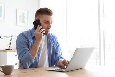 Photo of Young man talking on mobile phone while working with laptop at desk. Home office