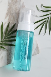 Photo of Bottle of face cleansing product and green leaves on white marble table, flat lay