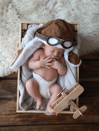 Cute newborn baby wearing aviator hat with toy sleeping in wooden crate, top view