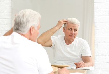 Senior man with hair loss problem looking in mirror indoors