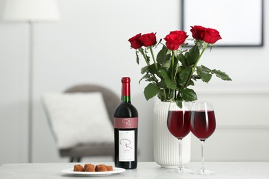 Photo of Bottle, glassesred wine, chocolate truffles and vase with roses on table in room. Romantic date