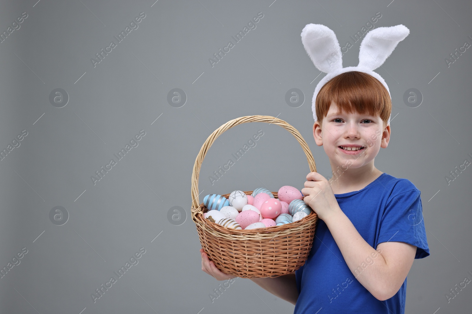 Photo of Easter celebration. Cute little boy with bunny ears and wicker basket full of painted eggs on grey background. Space for text