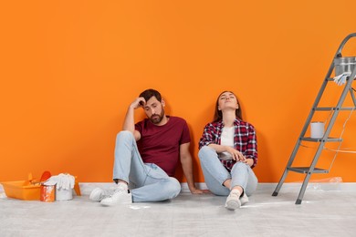 Photo of Tired designers sitting on floor with painting equipment near freshly painted orange wall indoors