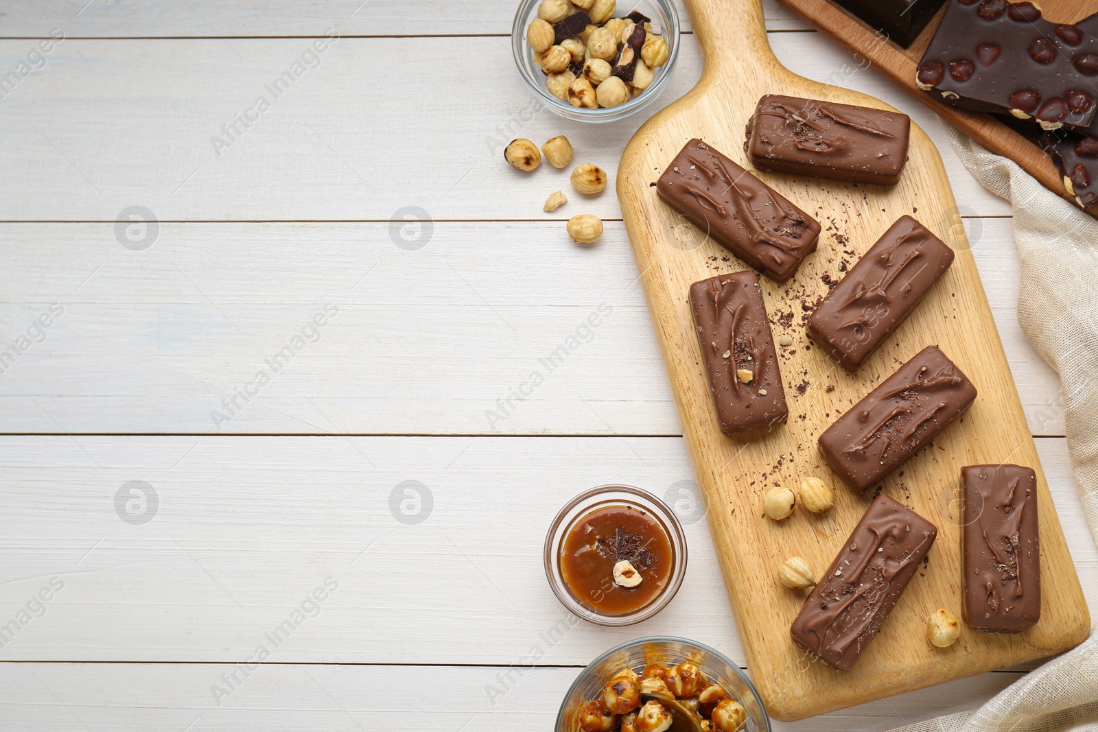 Photo of Delicious chocolate candy bars, caramel and nuts on white wooden table, flat lay. Space for text