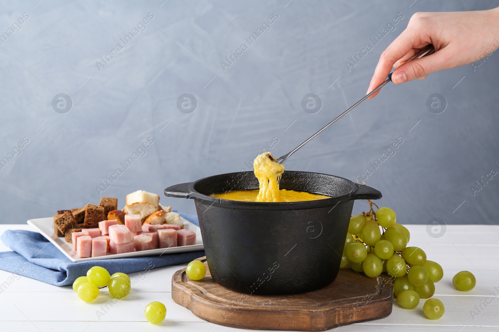 Photo of Woman dipping piece of bread into fondue pot with tasty melted cheese at white wooden table against gray background, closeup