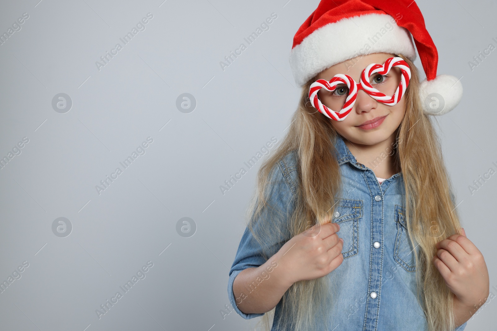 Photo of Girl wearing decorative Christmas eyeglasses in shape of hearts and Santa hat on light grey background, space for text