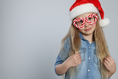 Girl wearing decorative Christmas eyeglasses in shape of hearts and Santa hat on light grey background, space for text