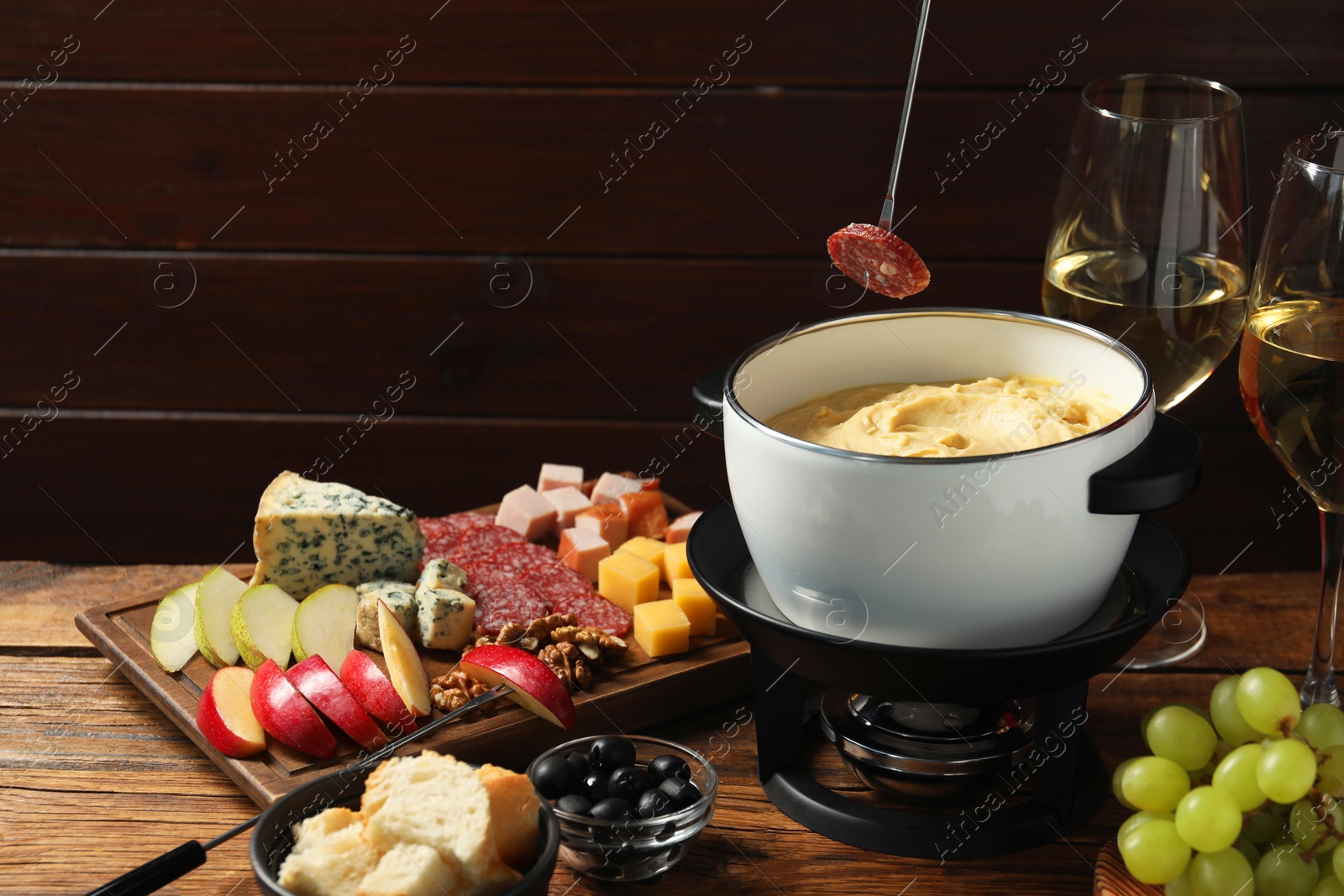 Photo of Dipping piece of sausage into fondue pot with melted cheese at wooden table with products, closeup