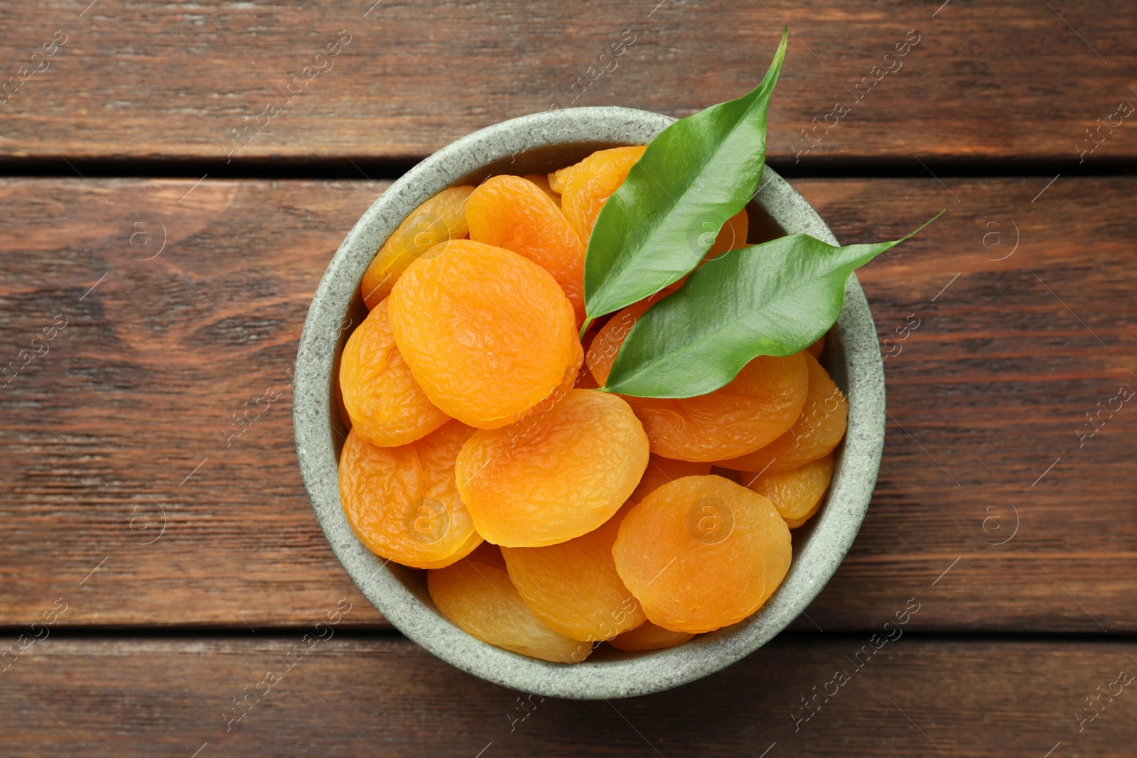 Photo of Bowl of tasty apricots and green leaves on wooden table, top view. Dried fruits