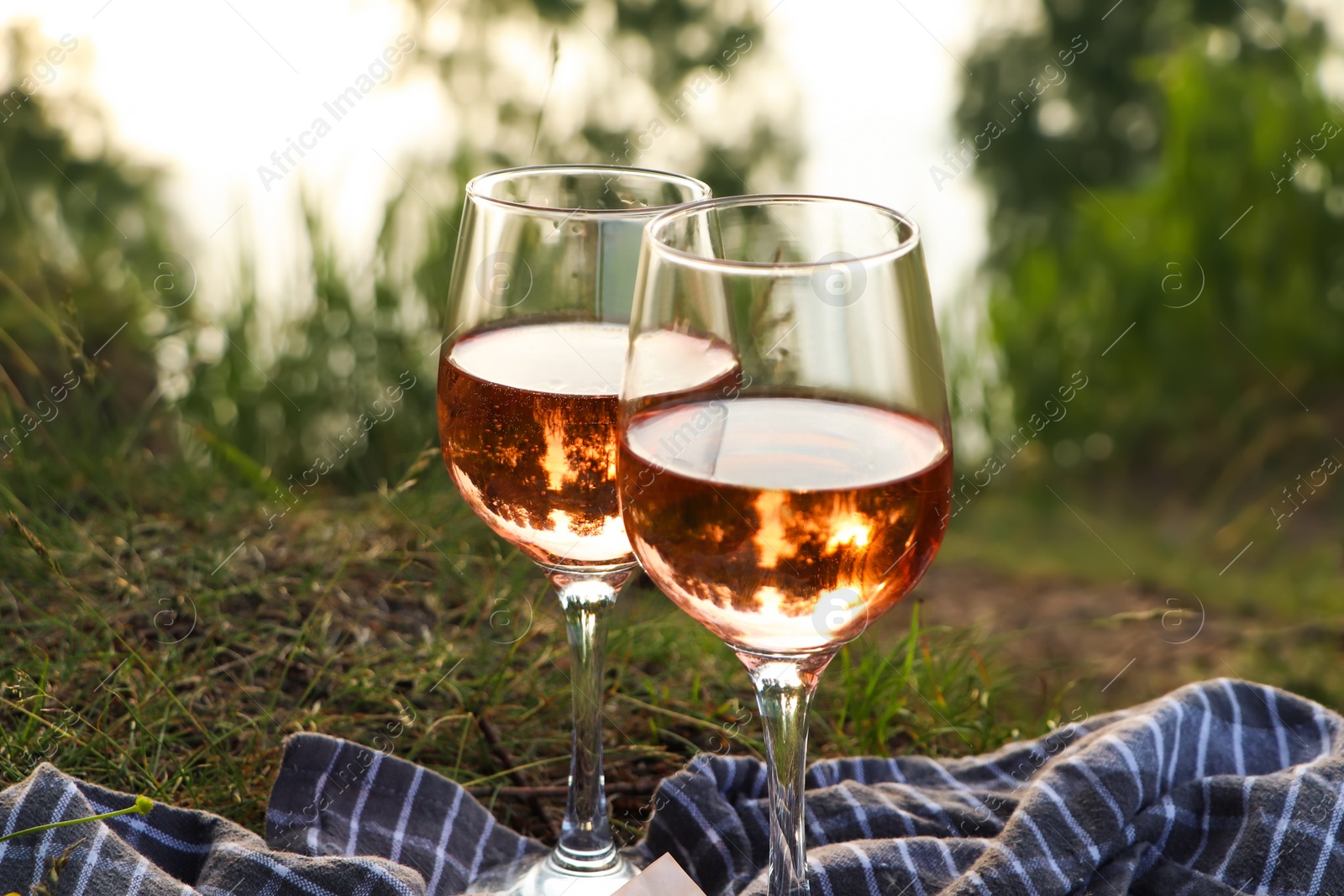 Photo of Glasses of delicious rose wine on picnic blanket near lake, closeup