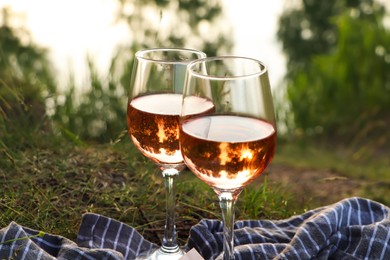 Glasses of delicious rose wine on picnic blanket near lake, closeup