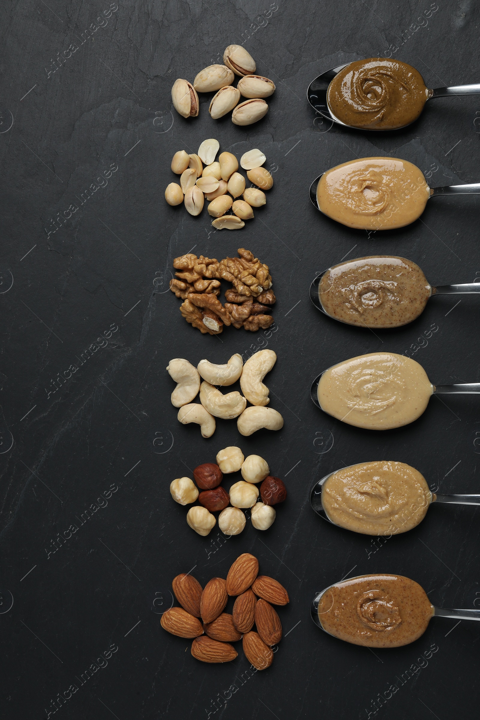 Photo of Tasty nut butters in spoons and raw nuts on dark gray textured table, flat lay