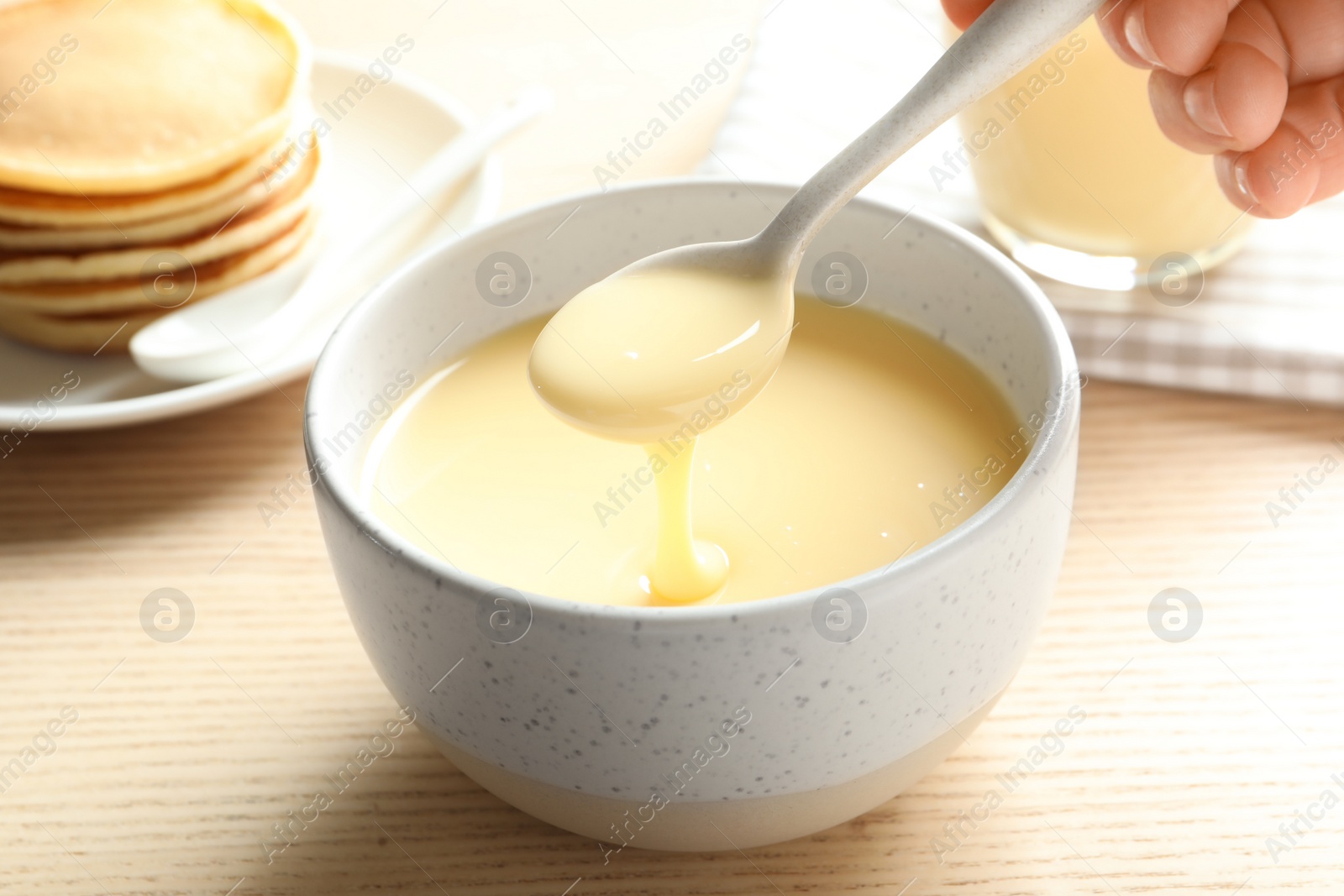 Photo of Person holding spoon with condensed milk over bowl on table, closeup. Dairy products