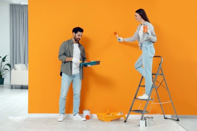 Photo of Woman painting orange wall and man holding container with roller indoors. Interior design
