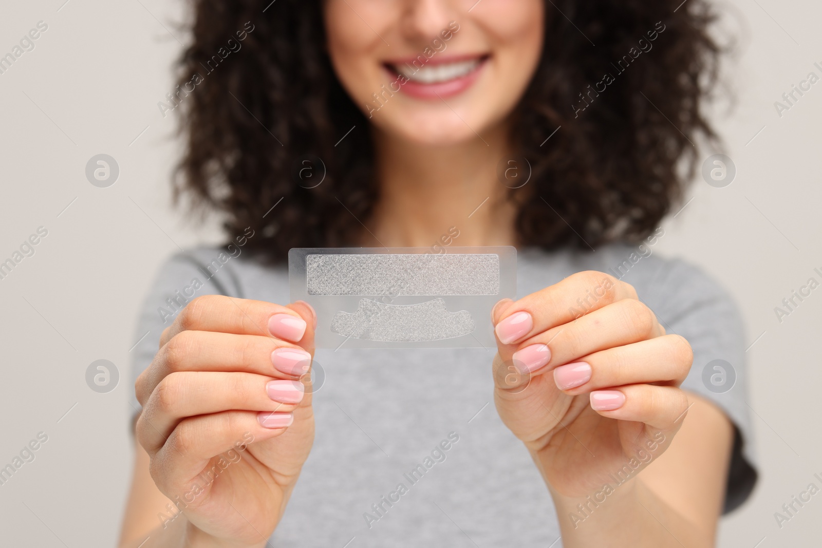 Photo of Young woman holding teeth whitening strips on light grey background, closeup