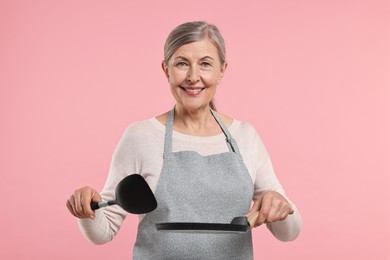 Happy housewife with turner and frying pan on pink background