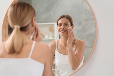 Beautiful woman removing makeup with cotton pad near mirror indoors