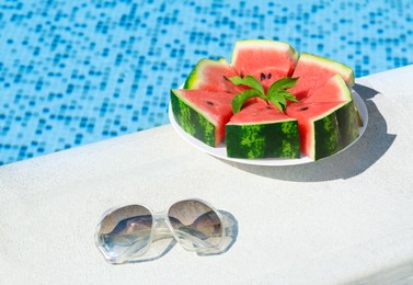 Photo of Slices of watermelon on white plate and sunglasses near swimming pool outdoors. Space for text
