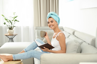 Pretty young woman with towel on head reading book and drinking coffee in living room