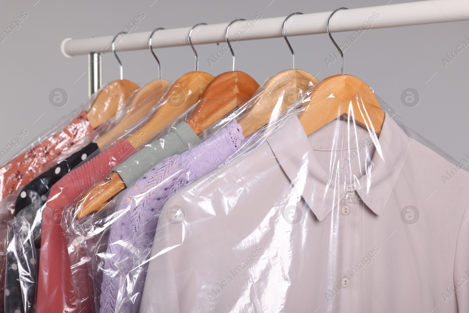 Photo of Dry-cleaning service. Many different clothes in plastic bags hanging on rack against grey background, closeup