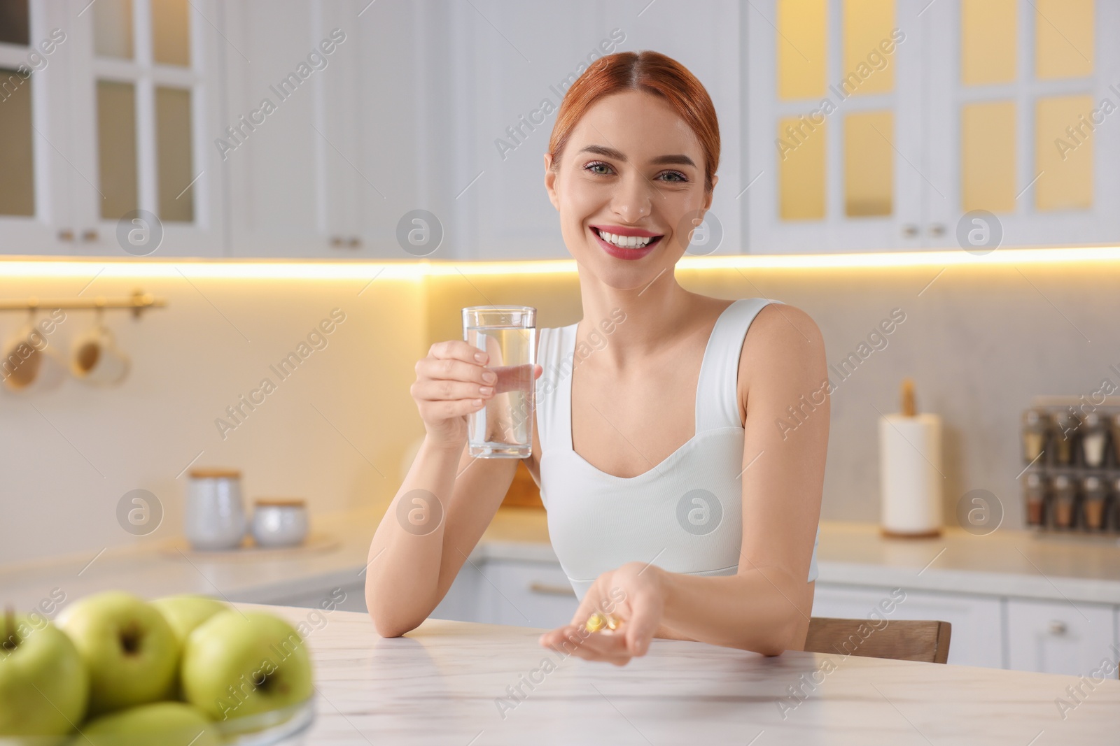 Photo of Happy young woman with glass of water and pills at table in kitchen. Weight loss