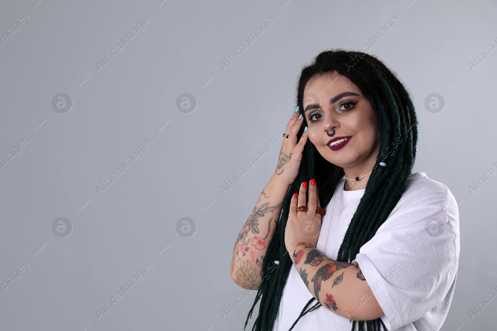 Photo of Beautiful young woman with tattoos on arms, nose piercing and dreadlocks against grey background. Space for text