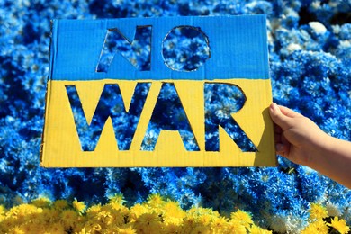 Photo of Woman holding poster in colors of Ukrainian flag with words No War and beautiful blue and yellow flowers on background, closeup