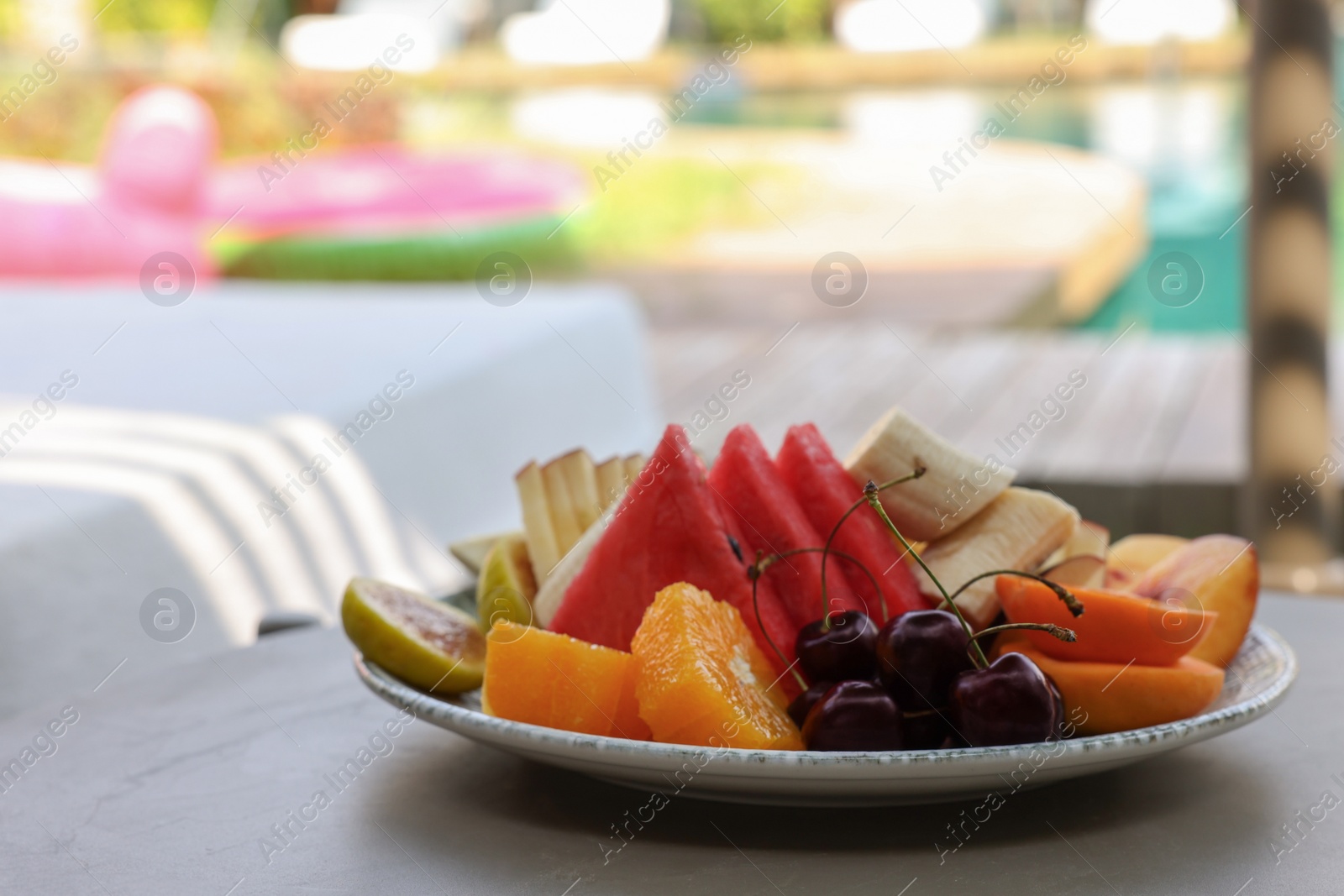 Photo of Plate with fresh fruits on table near sun lounger, space for text. Luxury resort with outdoor swimming pool