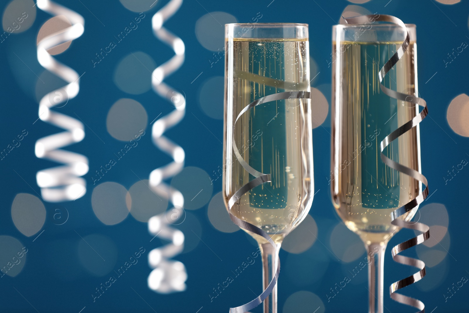 Photo of Glasses of champagne and serpentine streamers against blue background with blurred lights, closeup. Space for text