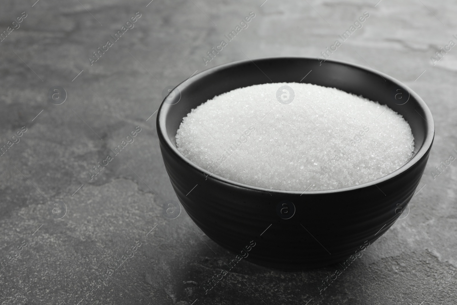 Photo of Granulated sugar in bowl on grey textured table, closeup. Space for text