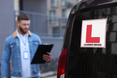 Instructor with clipboard near car outdoors, selective focus on L-plate. Driving school