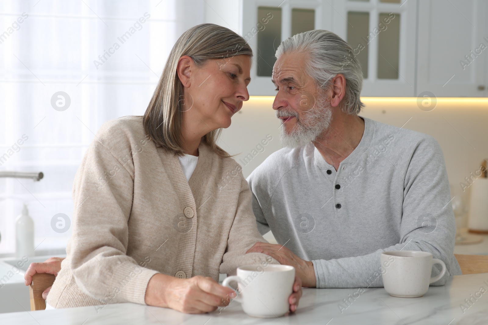 Photo of Affectionate senior couple with cups of drink at white marble table in kitchen