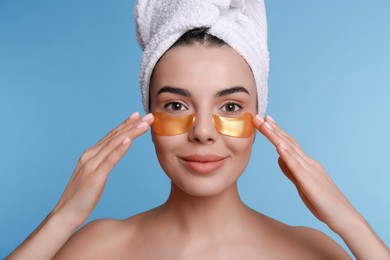 Beautiful young woman with under eye patches and hair wrapped in towel on light blue background