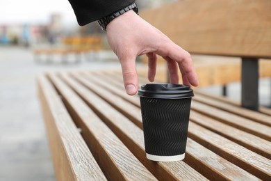 Man taking black paper cup from wooden bench outdoors, closeup. Space for text