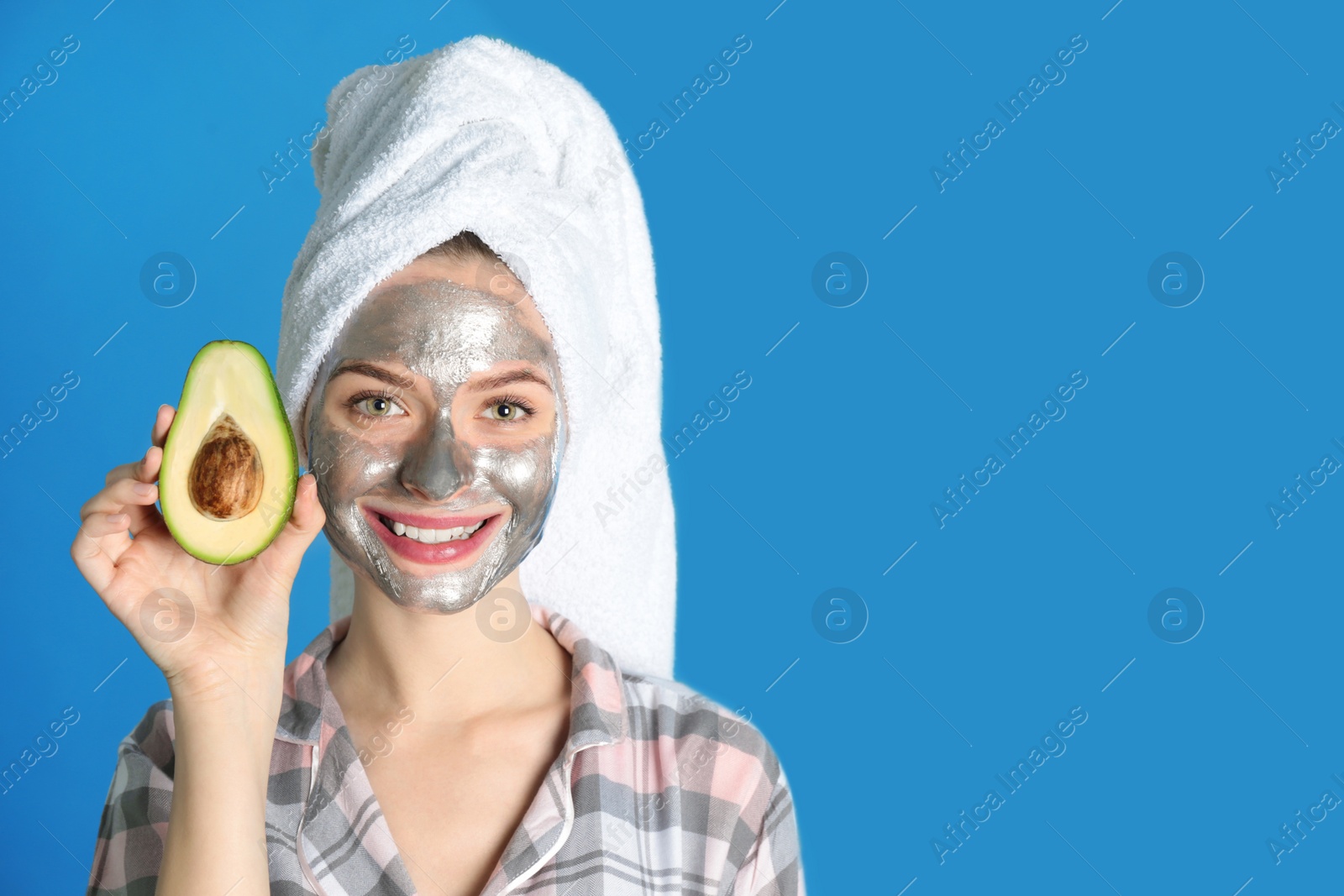 Photo of Young woman with cleansing mask on her face holding avocado against color background, space for text. Skin care