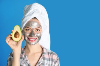 Photo of Young woman with cleansing mask on her face holding avocado against color background, space for text. Skin care