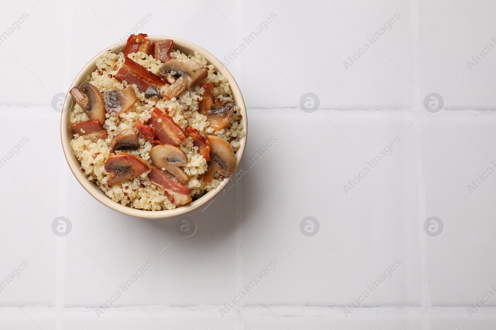 Photo of Tasty quinoa porridge with fried bacon and mushrooms in bowl on white tiled table, top view. Space for text