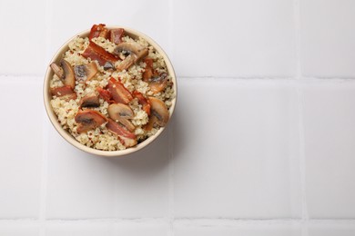 Photo of Tasty quinoa porridge with fried bacon and mushrooms in bowl on white tiled table, top view. Space for text