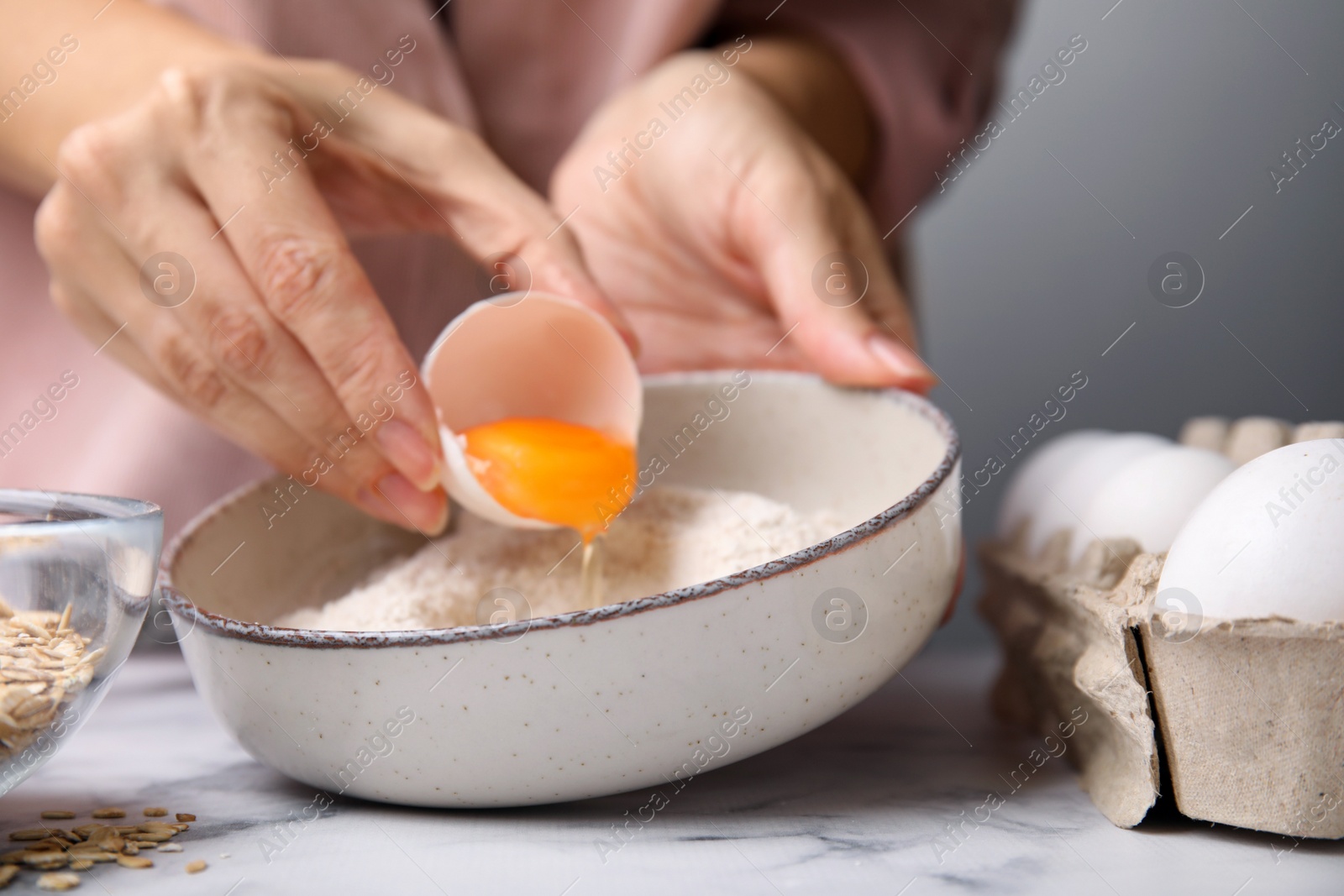 Photo of Woman adding egg to bowl with flour at white marble table in kitchen, closeup. Cooking oatmeal dough