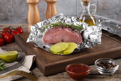 Photo of Aluminum foil with raw meat, rosemary, lime and spices on wooden table