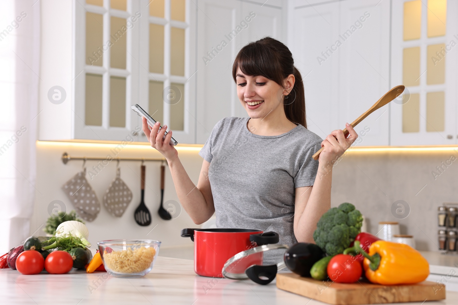 Photo of Happy young housewife with spoon using smartphone while cooking at white marble table in kitchen