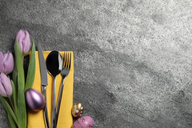 Photo of Cutlery set, painted eggs and beautiful flowers on grey table, flat lay with space for text. Easter celebration