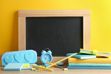 Different school stationery and small blank chalkboard on table near yellow wall. Space for text