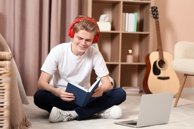 Photo of Online learning. Smiling teenage boy in headphones reading book near laptop at home