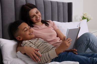 Photo of Mother with her African American son reading book on bed at home. International family